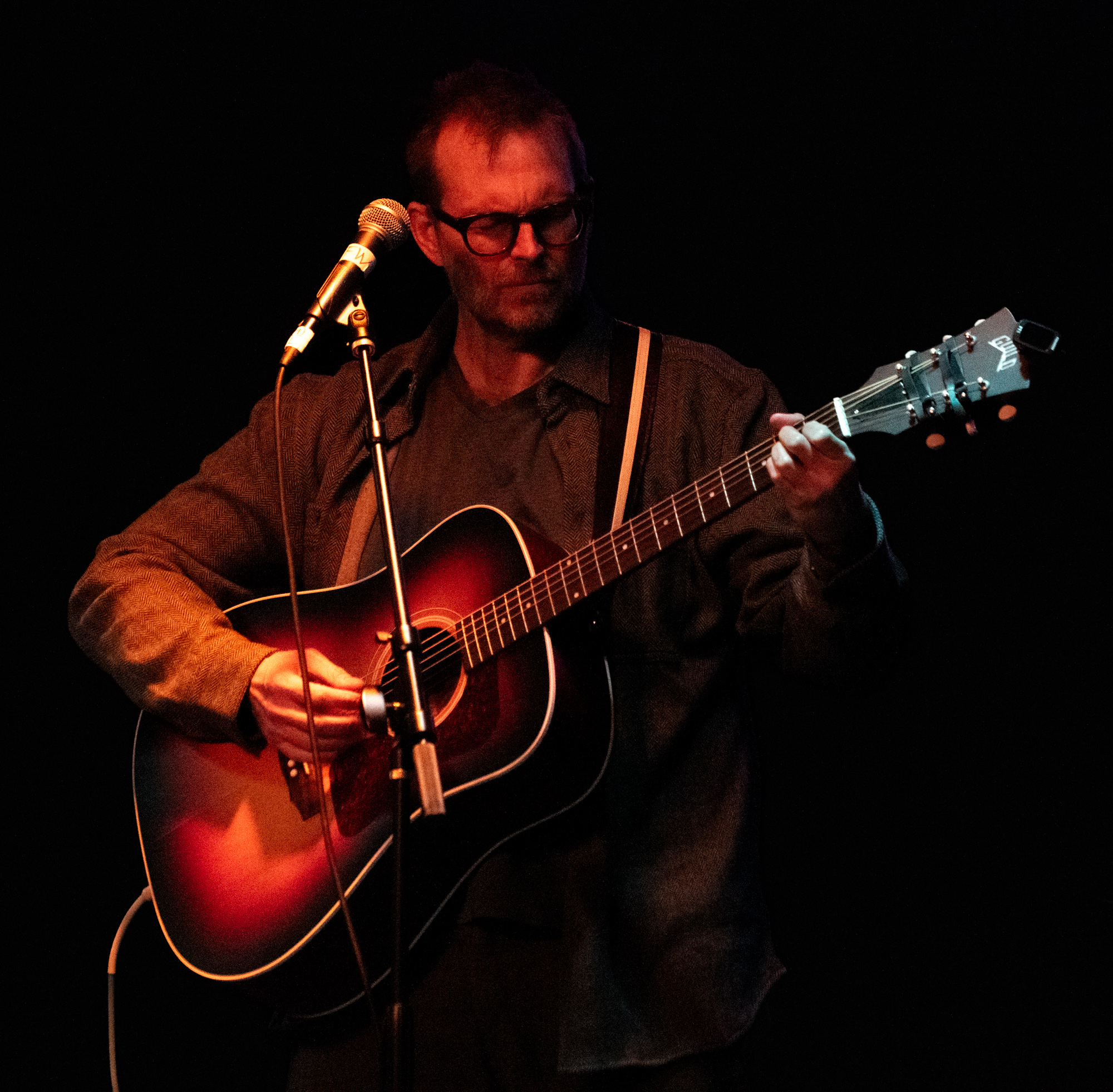 Ian Felton looking down at his acoustic guitar, warm red stage light on his face