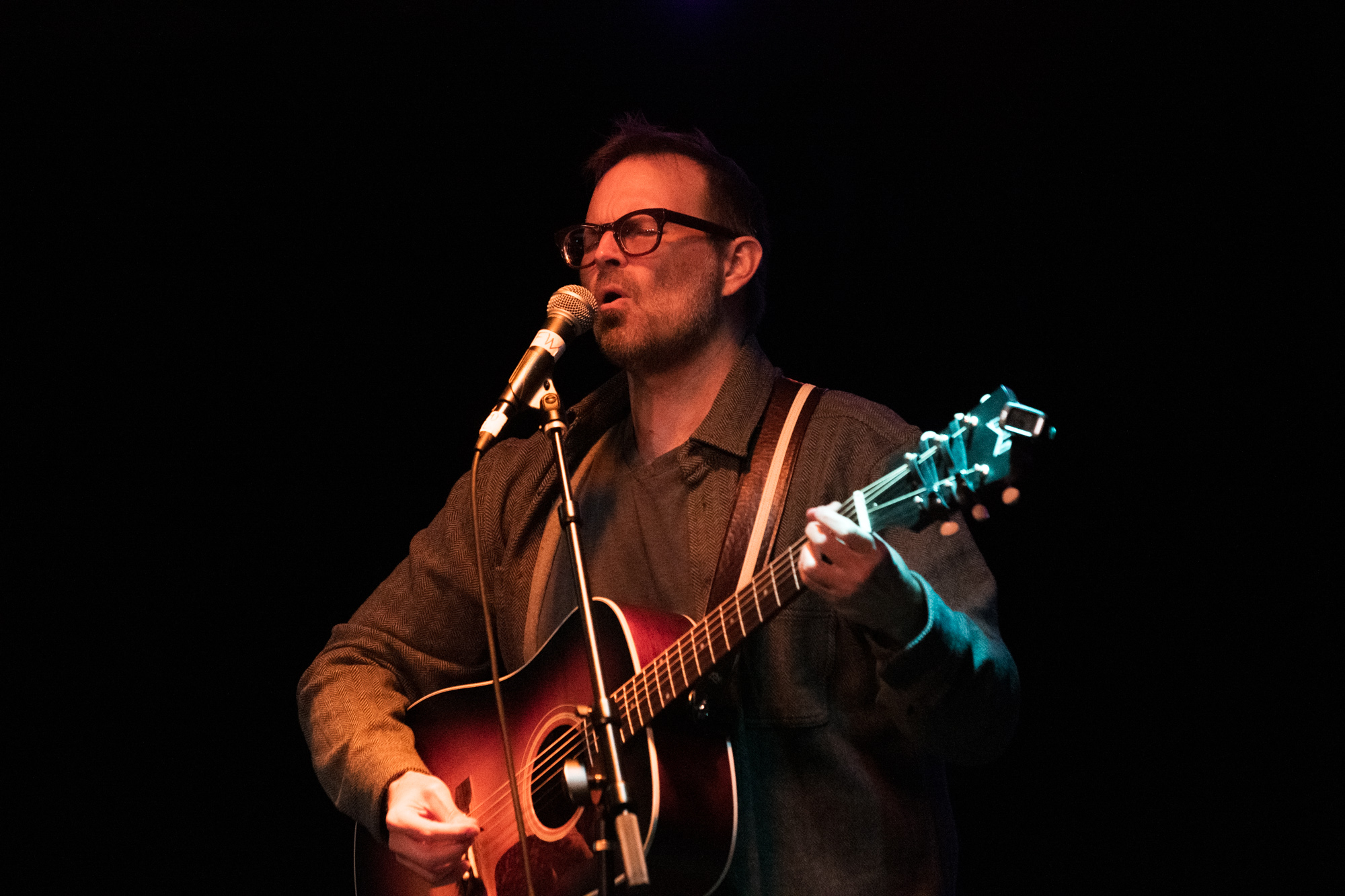 Ian Felton mid-vocal at the microphone, eyes nearly closed, his Guild guitar caught between warm and teal stage light
